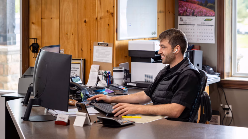 Man working at a desk in a Buckeye Metal Sales office, with computer monitor, paperwork, and filing materials visible.