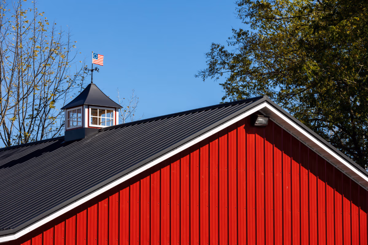 Red metal barn with black metal roof and white trim, topped with a cupola and weathervane against a clear blue sky