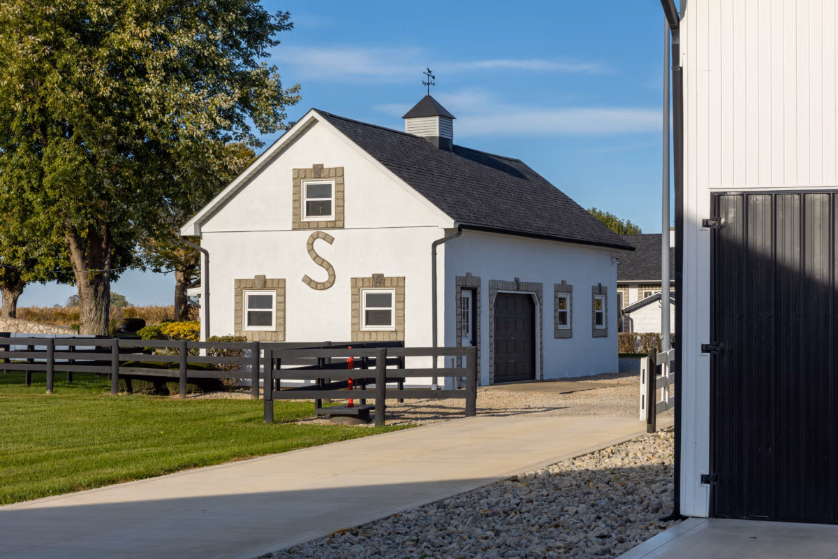 White barn with attached structure featuring a cupola with weathervane on the roof, metal roofing and siding, with wooden rail fence and green lawn