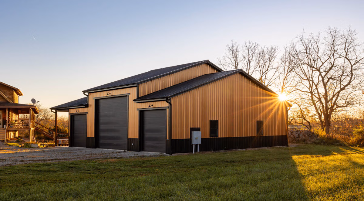 Metal rectangular pole barn with dark metal roof, photographed at sunset with bare trees visible in the background.