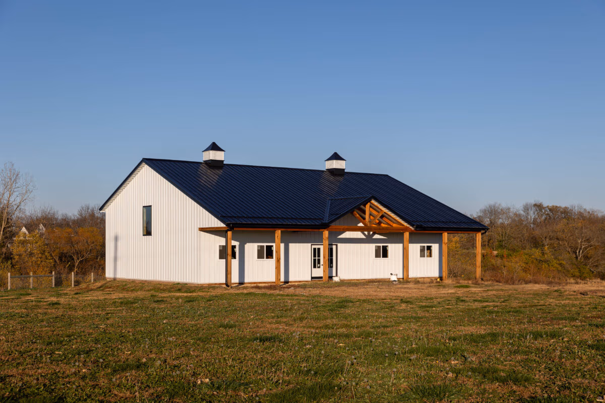 White pole barn with tan metal siding, dark metal roof, and cupolas, situated in open field