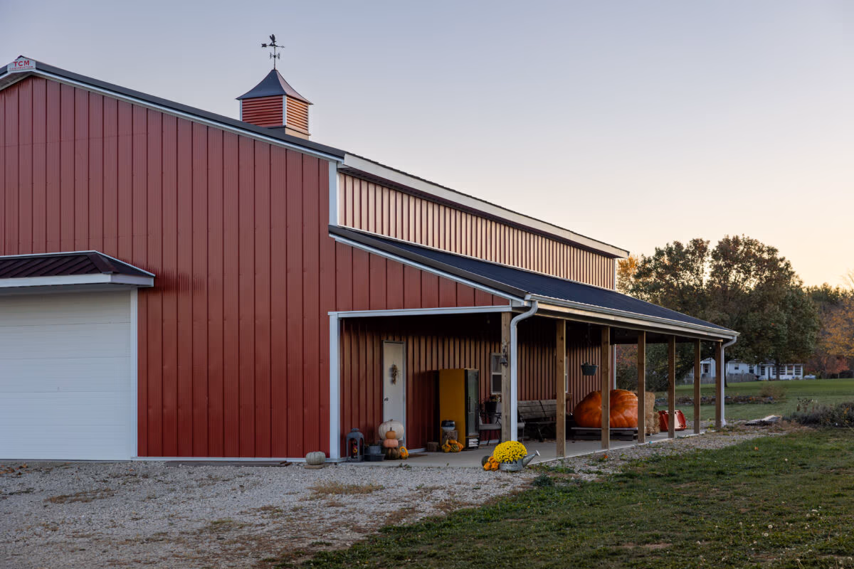 A residential pole barn with red corrugated metal siding, white trim, gabled roof with cupola, and a covered front porch entry.