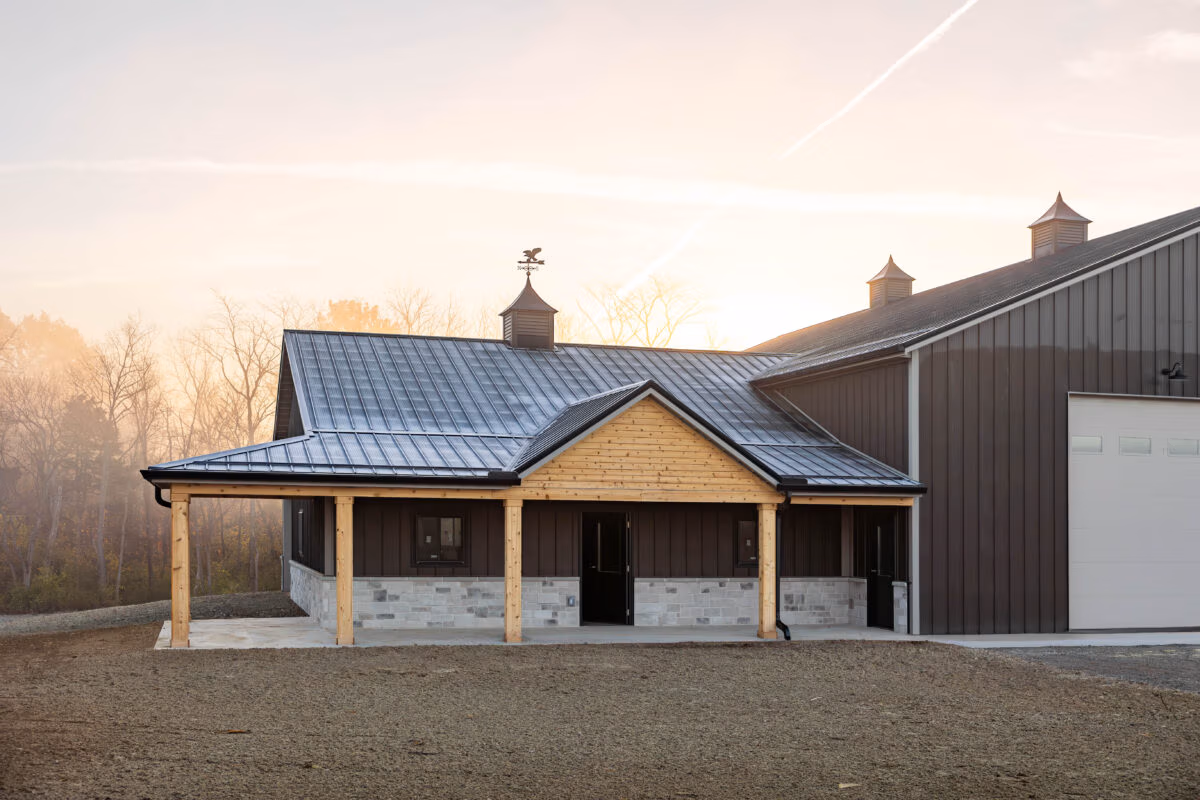 Tan pole barn with standing seam metal roof and small cupola with weathervane on ridge, dark metal siding on adjacent structure