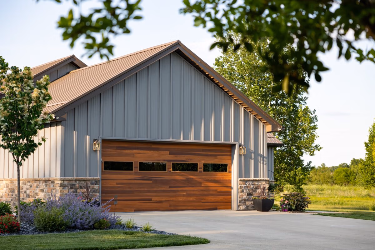 Residential garage with vertical metal siding, metal roof with overhang, and wooden garage door, surrounded by landscaping and trees