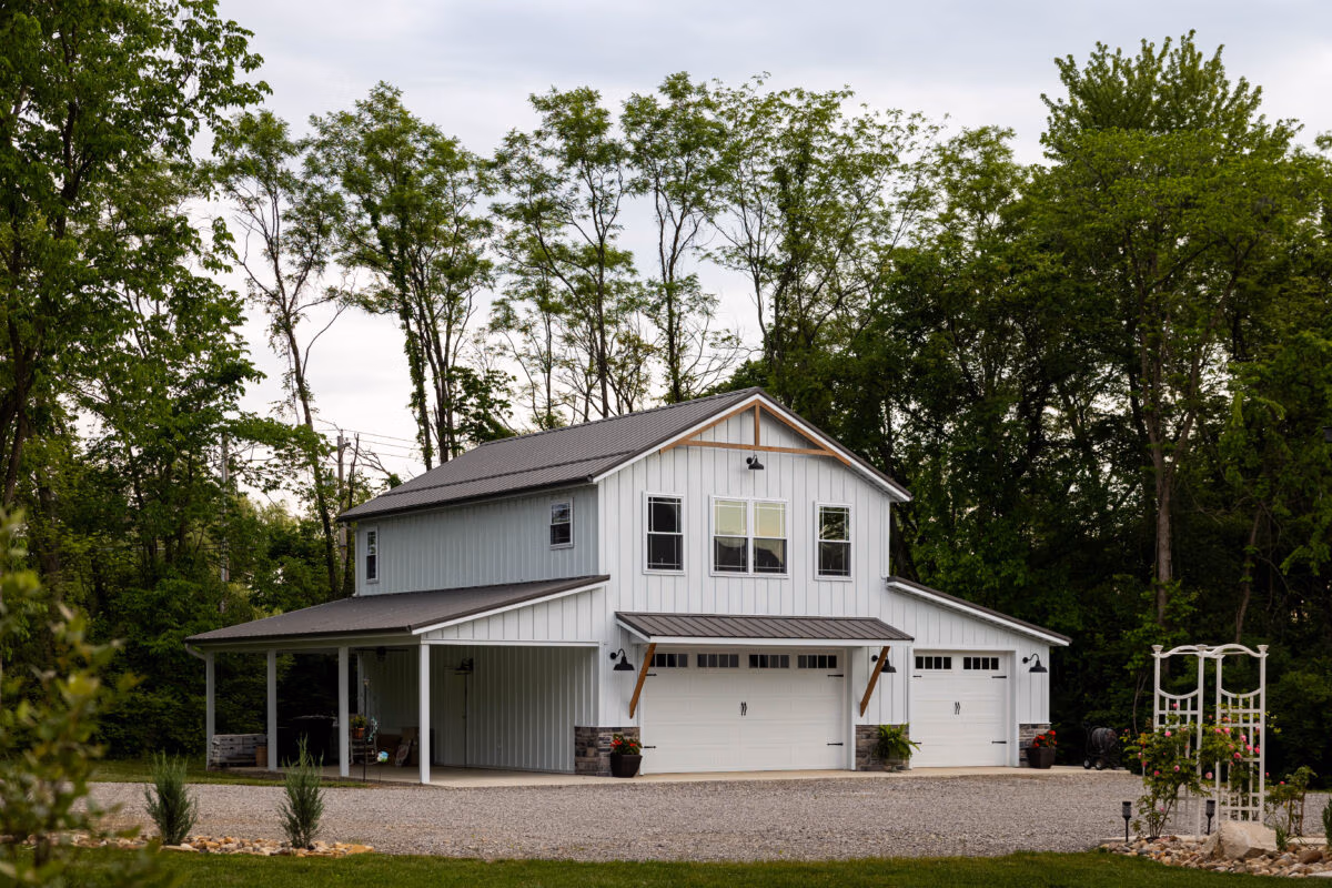 Two-story residential home with white metal siding, metal roof, attached garage, and wraparound porch surrounded by mature trees