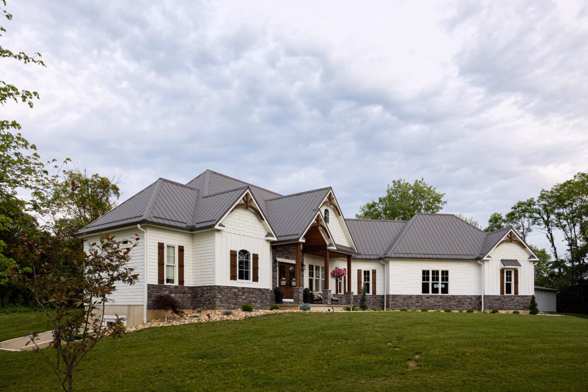 Two-story residential home with dark metal roof, cream-colored siding, and manicured lawn surrounded by mature trees