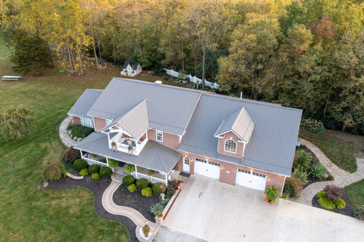 Aerial view of a residential home with a gray metal roof and brick siding, surrounded by mature trees and landscaped driveway.