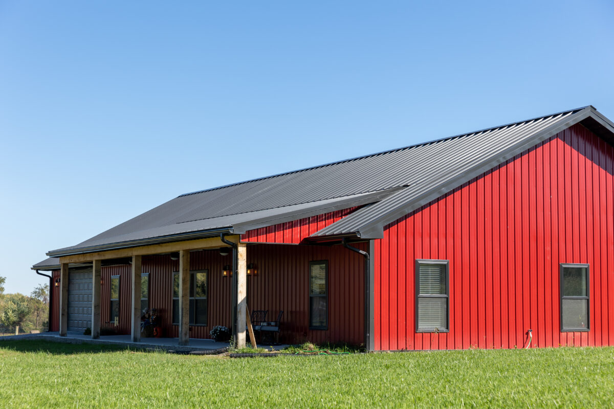Modern barn with bright red metal siding and dark metal roof, featuring covered porch area on grass field